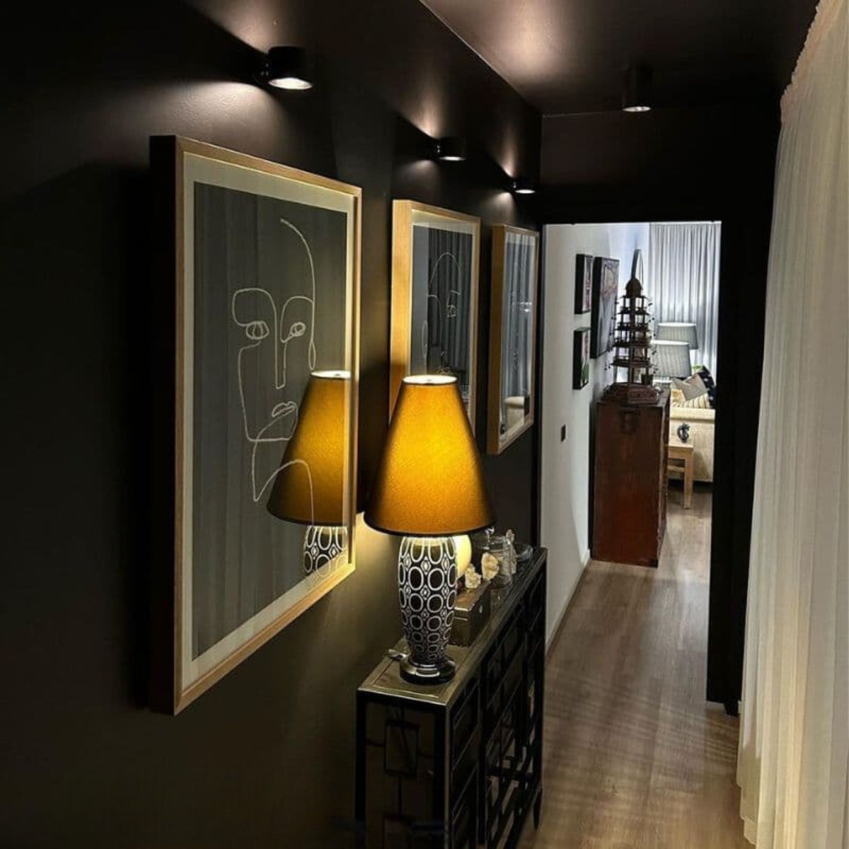 Several black wall lights in a dark hallway, shining down on framed line-drawing art above a console table and lamp. 10