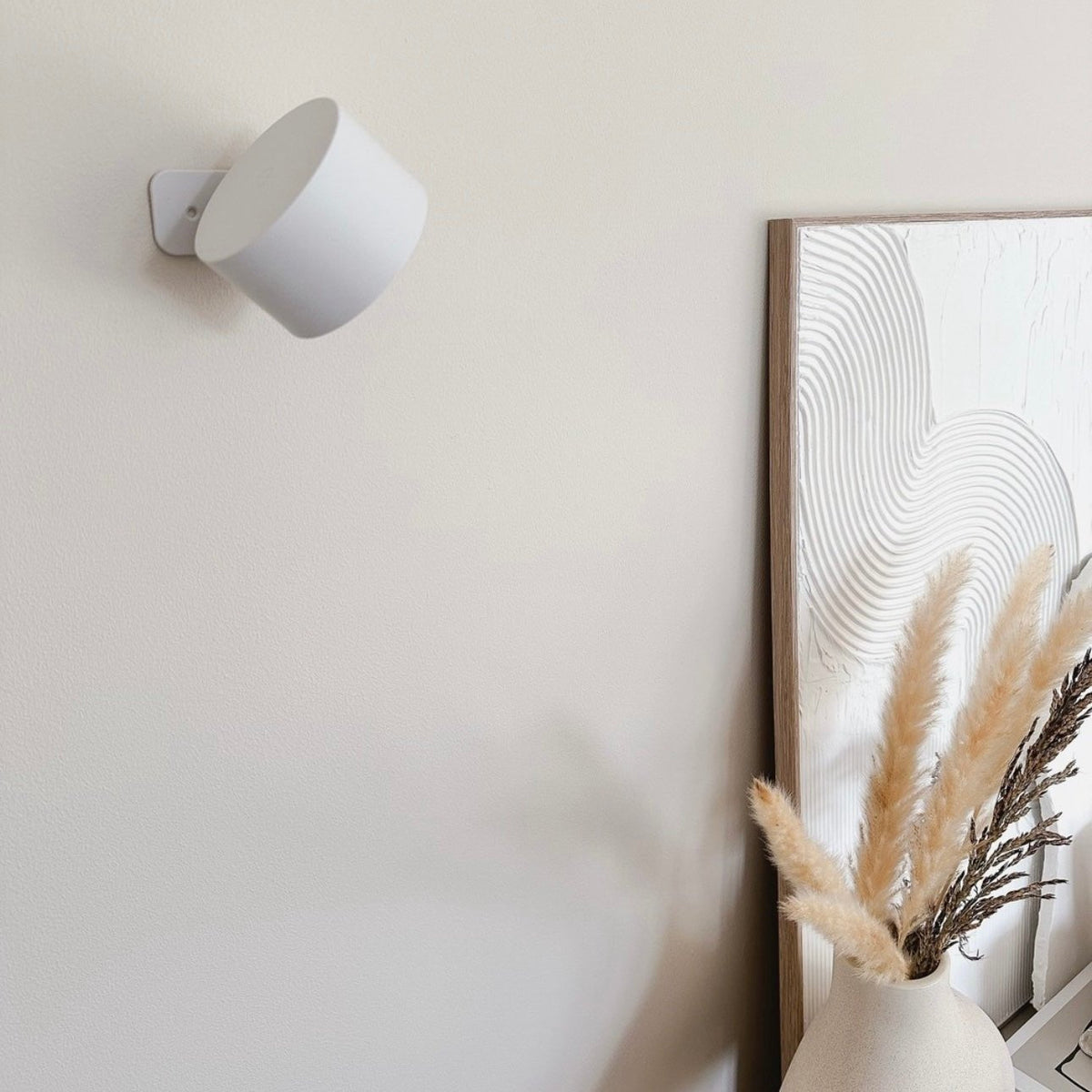 White wall light switched off on a cream wall, tilted away, next to a framed textured artwork and dried pampas grass. 17