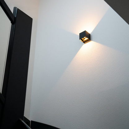 Black cube wall lamp glowing warmly on a white textured stairwell wall, seen from below beside a dark doorframe. 2