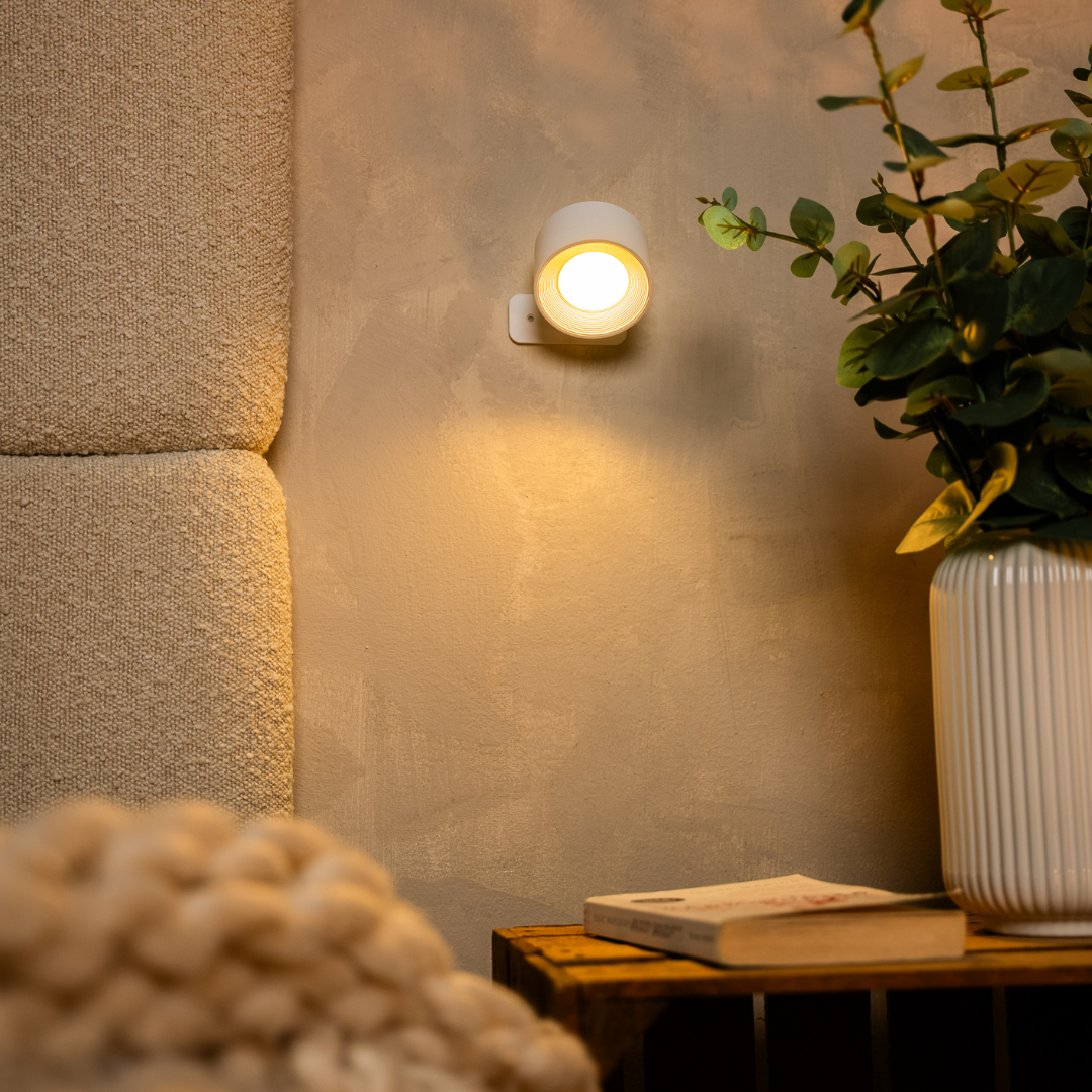 White wall light switched on against a textured wall, beside a ribbed white vase of eucalyptus and a stack of books. 12
