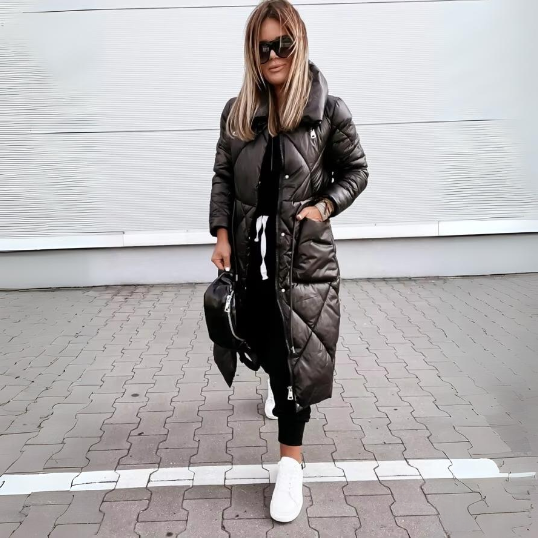 Woman with blonde hair wears the black quilted long coat zipped up, one hand in a pocket, with white trainers. 5