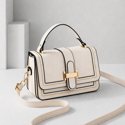 Cream flap bag with top handle, gold clasp and contrast stitching, shown at an angle with its long strap curled on a pale studio set. 1