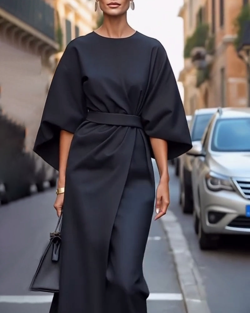 Black wrap-front dress with flowing sleeves and tied waist belt, worn by a model with earrings, a gold watch and a small black handbag. 6