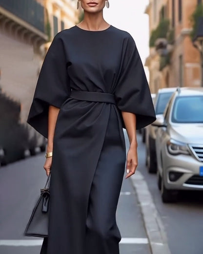 Black wrap-front dress with flowing sleeves and tied waist belt, worn by a model with earrings, a gold watch and a small black handbag. 6