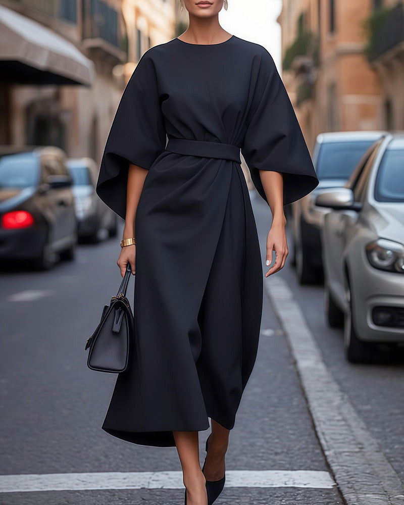Full-length view of a model walking down a city street in the black belted midi dress, carrying a black handbag. 1
