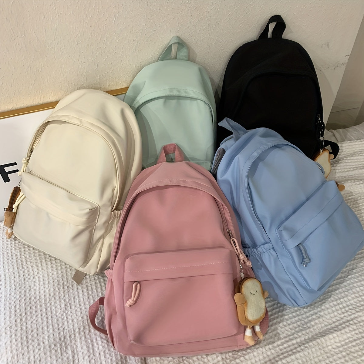 Five backpacks in cream, mint, black, pink and light blue lying on a bed, two with small toast-shaped charms attached. 8