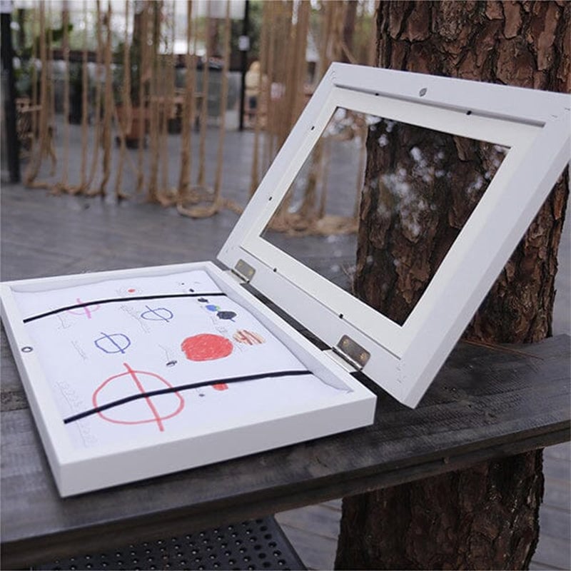 White frame opened flat on an outdoor table, a child's drawing held inside by two black elastic bands. 20