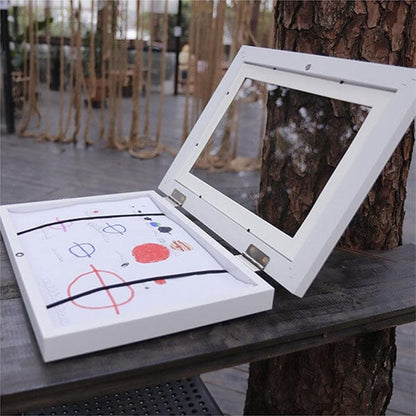 White frame opened flat on an outdoor table, a child's drawing held inside by two black elastic bands. 20
