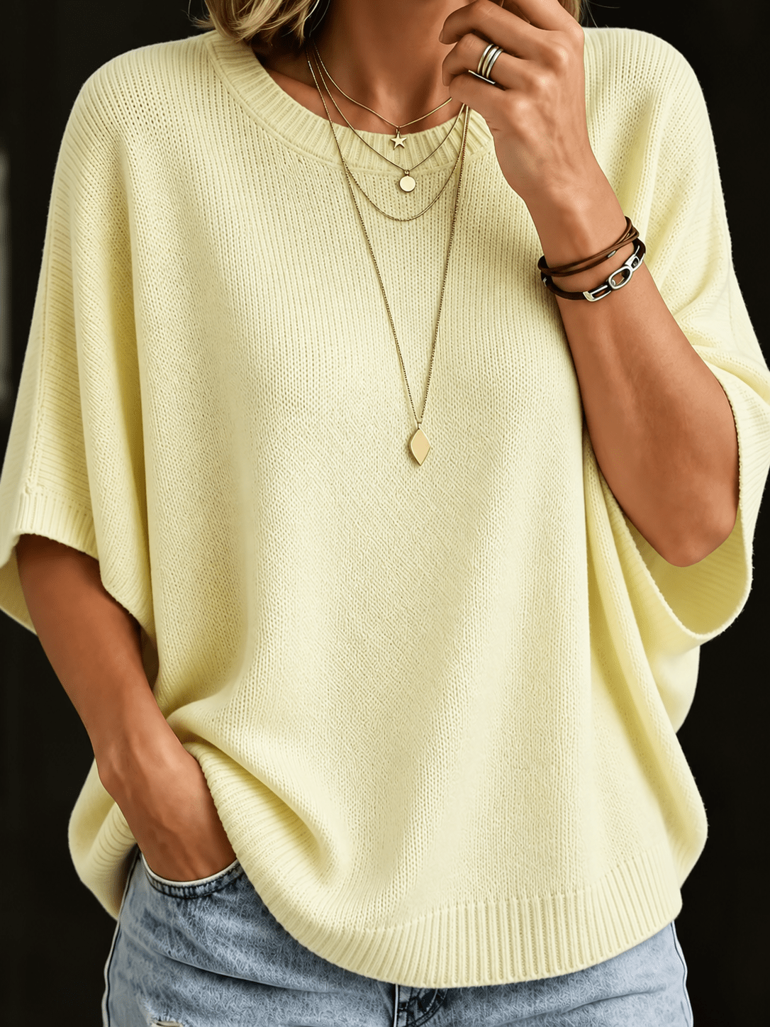 Close-up front view of the pale yellow batwing knit top with ribbed neckline and layered gold necklaces. 4