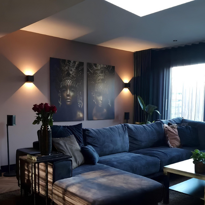 Two black cube wall lamps lighting a living room wall around two portrait canvases, above a blue corner sofa. 9