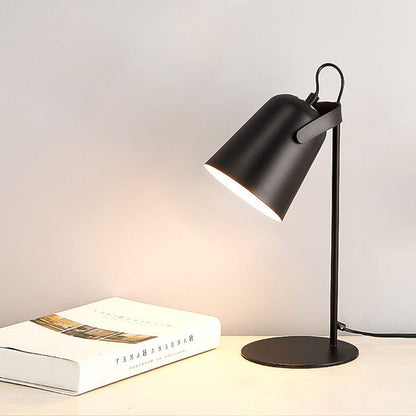 Black version of the lamp lit on a pale desk beside an open hardback book, against a soft grey wall. 12