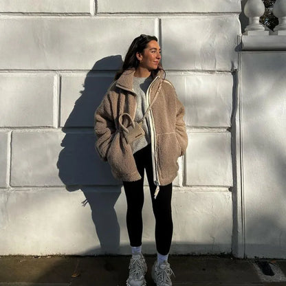 Woman in a beige fleece coat with hands in pockets, standing against a white wall in sunlight 4
