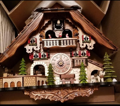 Chalet cuckoo clock sitting unhung inside its open cardboard box, showing the carved base, fir trees, balcony figures and a dog. 14