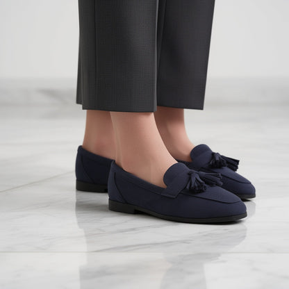Navy tasselled loafers worn with grey trousers on a white marble floor, side view 1