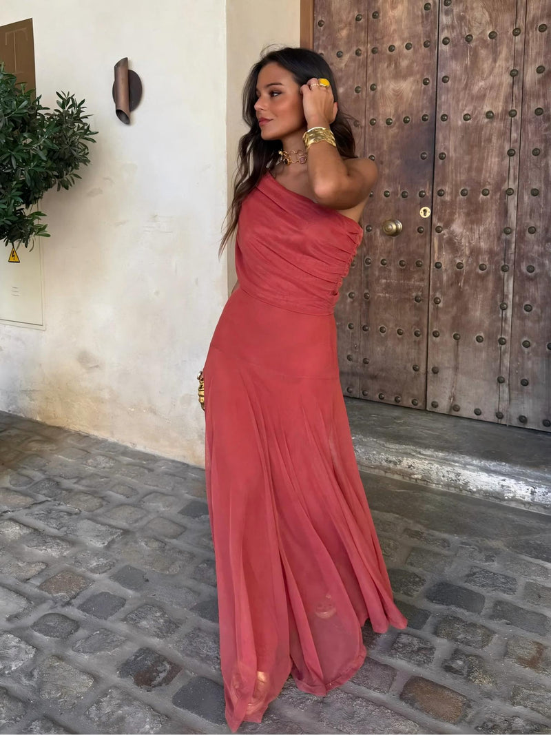 Model in terracotta one-shoulder mesh dress, hand in hair, standing before a studded wooden door 4