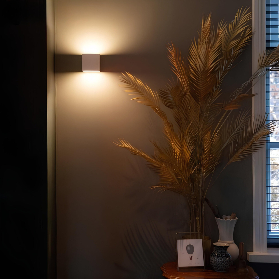 White cube wall lamp lit on a dark wall above a table, with golden palm fronds and small vases nearby. 4