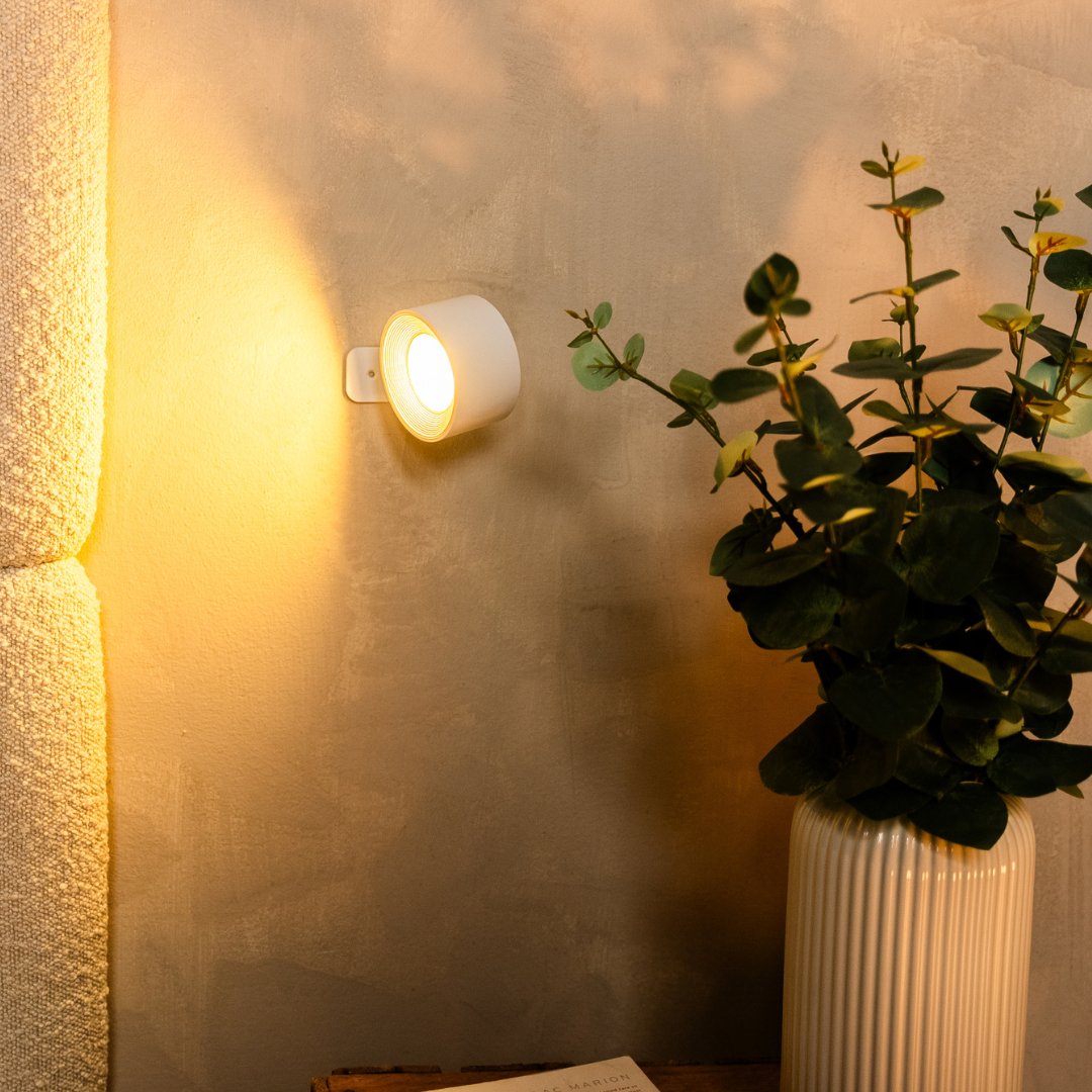White wall light glowing warmly on a plaster wall, with its mounting plate visible beside a vase of eucalyptus. 14