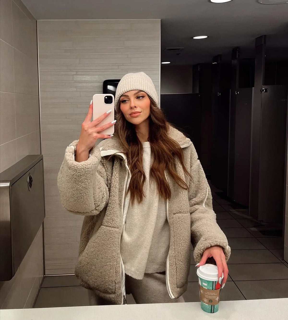 Woman in a beige fleece coat and cream beanie taking a mirror selfie, holding a takeaway cup 5