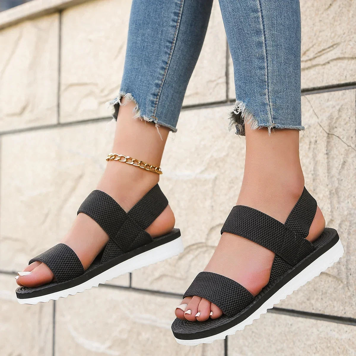 Black elastic-strap sandals worn with cropped jeans, feet raised against a pale stone wall 5