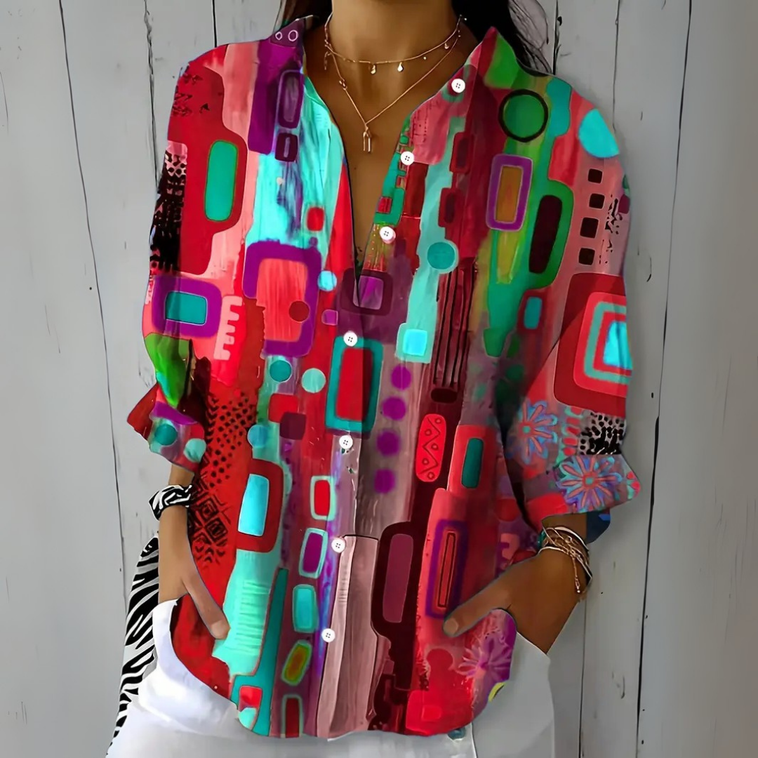 Model wearing the blouse in a red, turquoise and purple colourway with the abstract print, hands tucked into trouser pockets. 4