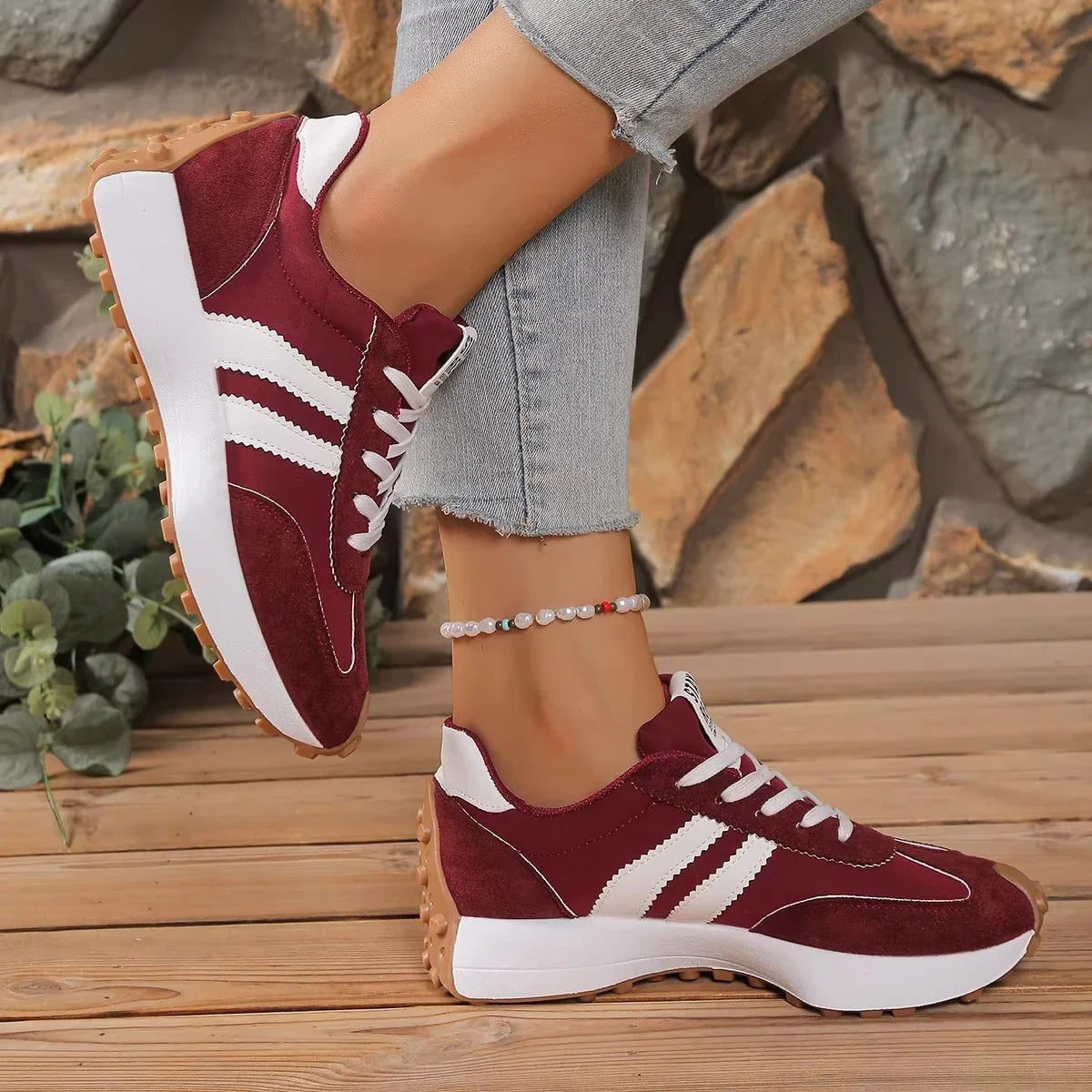 Burgundy trainers with white stripes worn with frayed jeans, one foot raised, on wooden decking by stone 5
