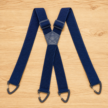 Navy blue X-back suspenders laid flat on wood, with slide adjusters near the centre and dark triangular clips at each end. 12