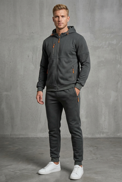 Man in dark grey hooded tracksuit with orange zip pulls, one hand in trouser pocket 4