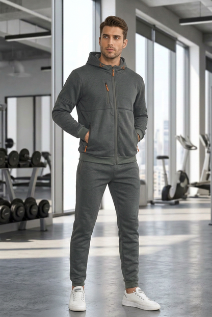Model in a grey zip-up hooded tracksuit with orange zip pulls, standing in a gym 1