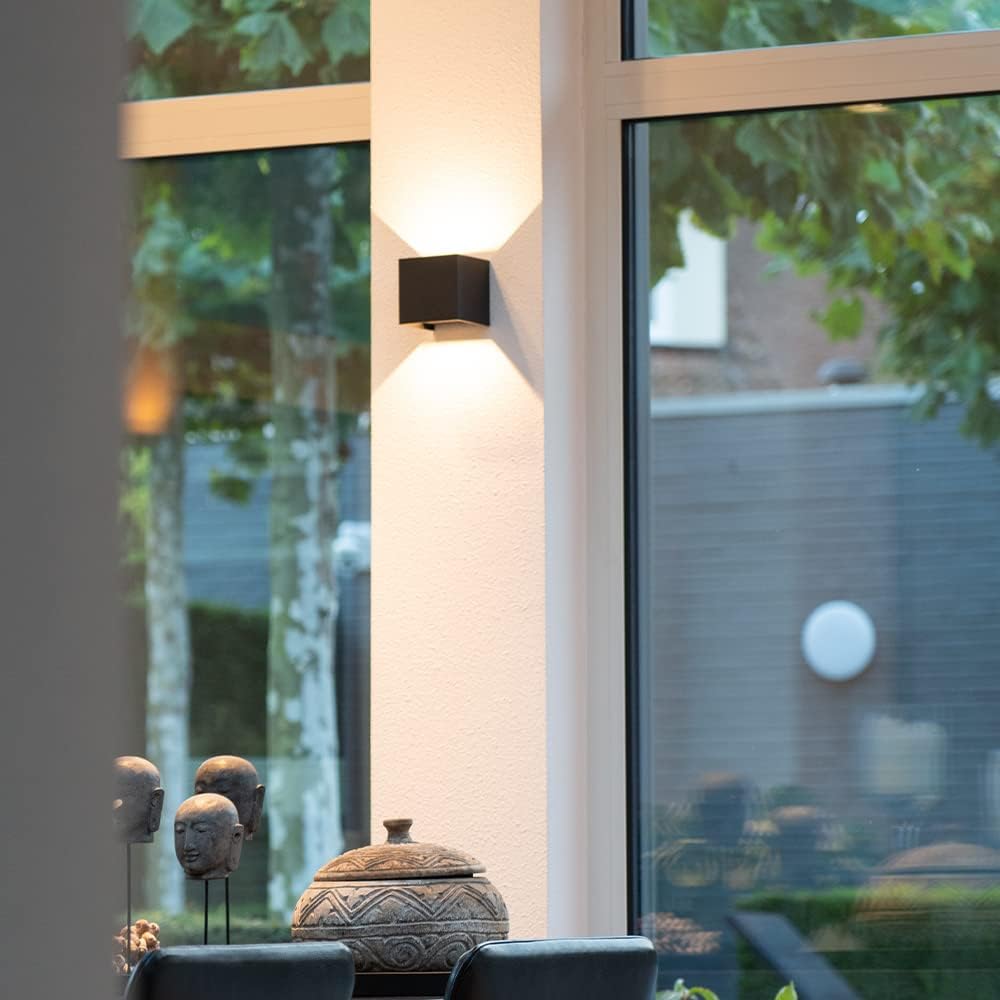 Black cube wall lamp on a pale pillar between large windows, with decorative stone heads and a lidded bowl below. 8
