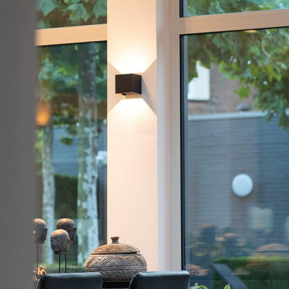 Black cube wall lamp on a pale pillar between large windows, with decorative stone heads and a lidded bowl below. 8