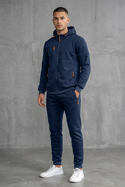 Man in navy hooded tracksuit with orange zip pulls, front view against a concrete wall 2