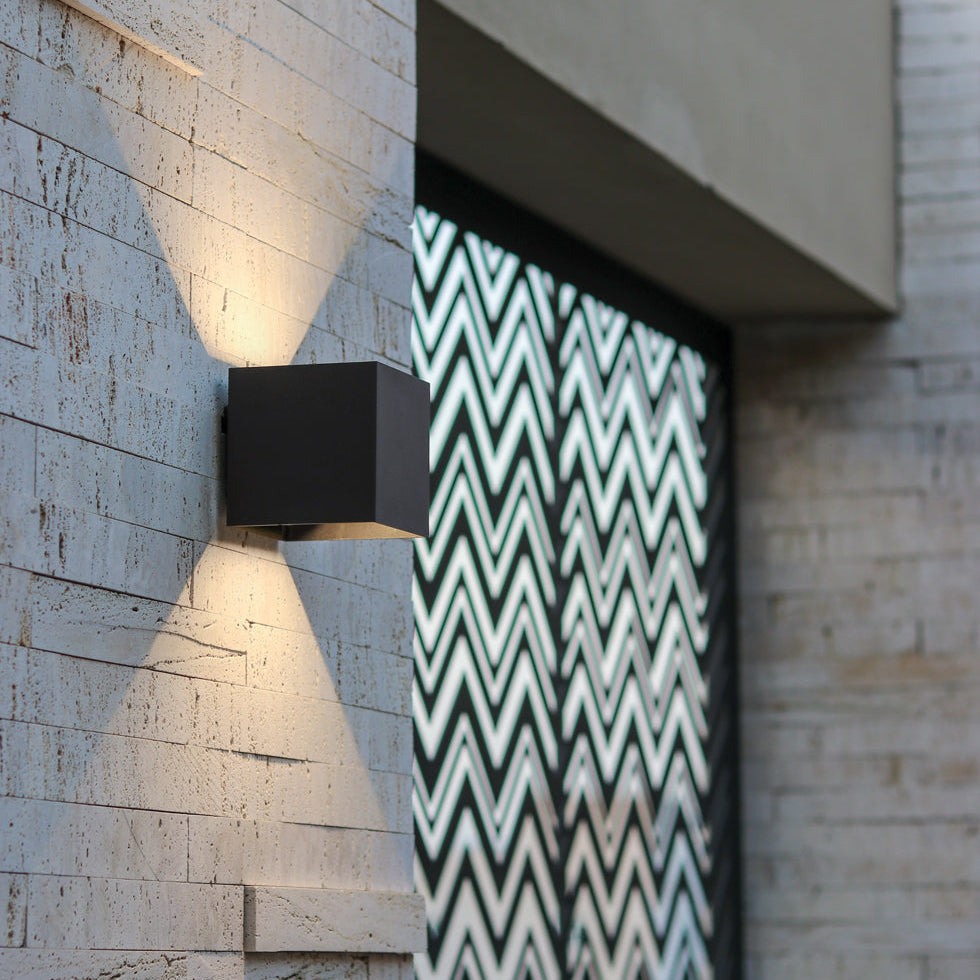 Close-up of a black cube wall lamp on a stone-clad exterior wall, sending light up and down beside a chevron-patterned panel. 11