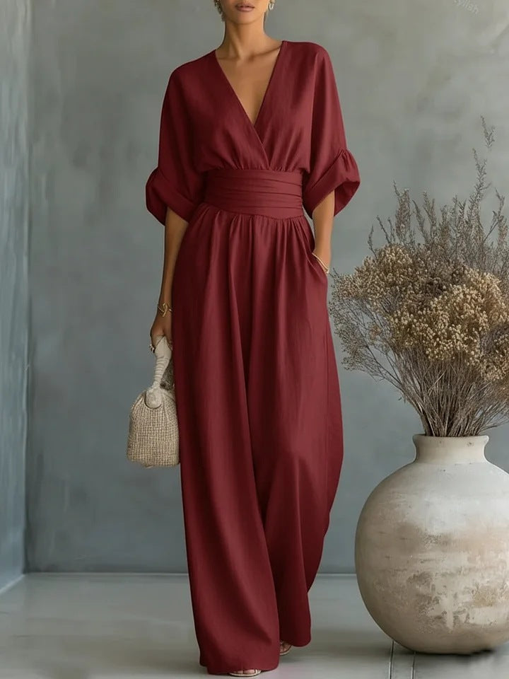 The jumpsuit in deep burgundy red, worn with rolled half sleeves and a woven handbag beside a stone vase. 4