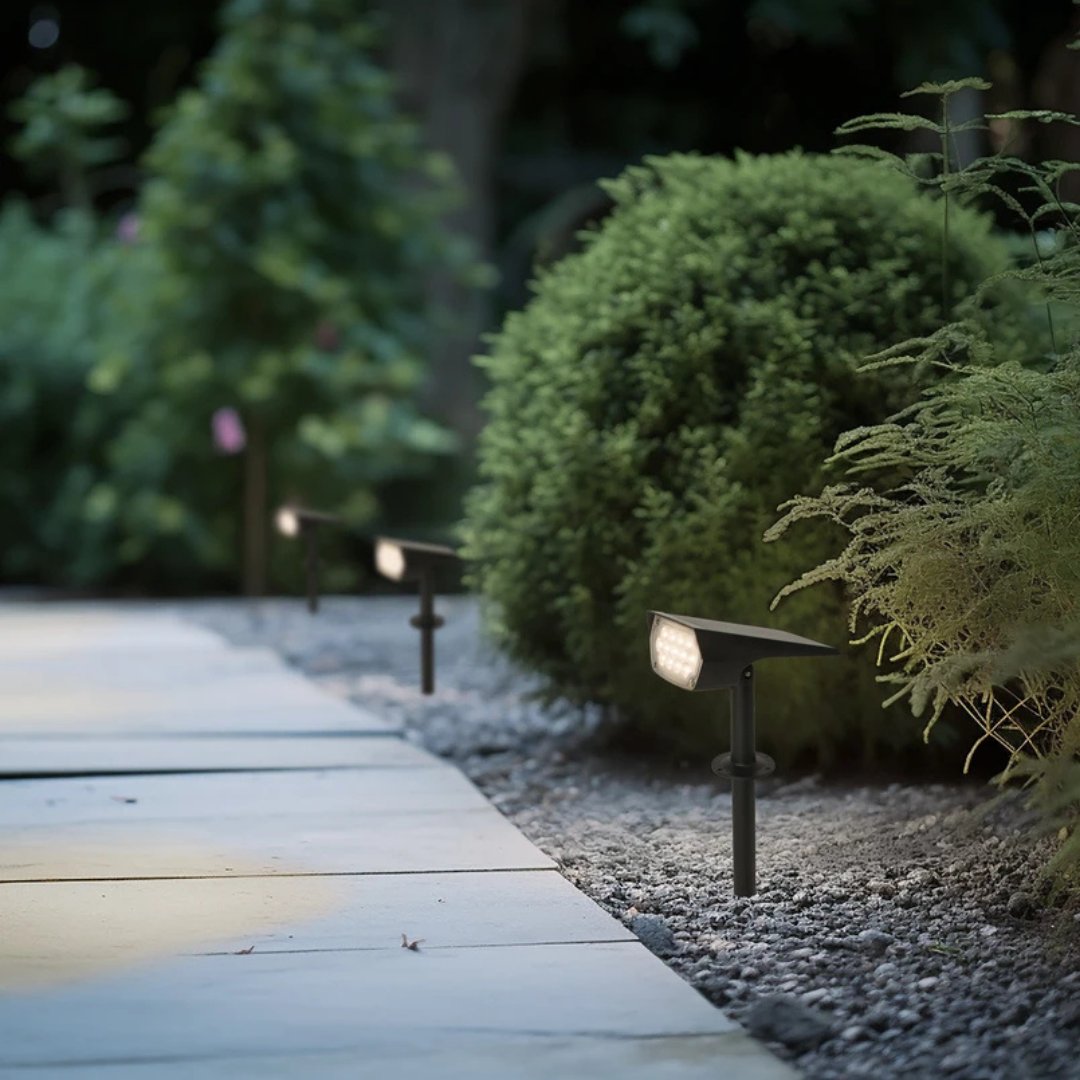 Black spotlights on stakes glowing warm white along a gravel border beside a paved garden path at dusk. 1