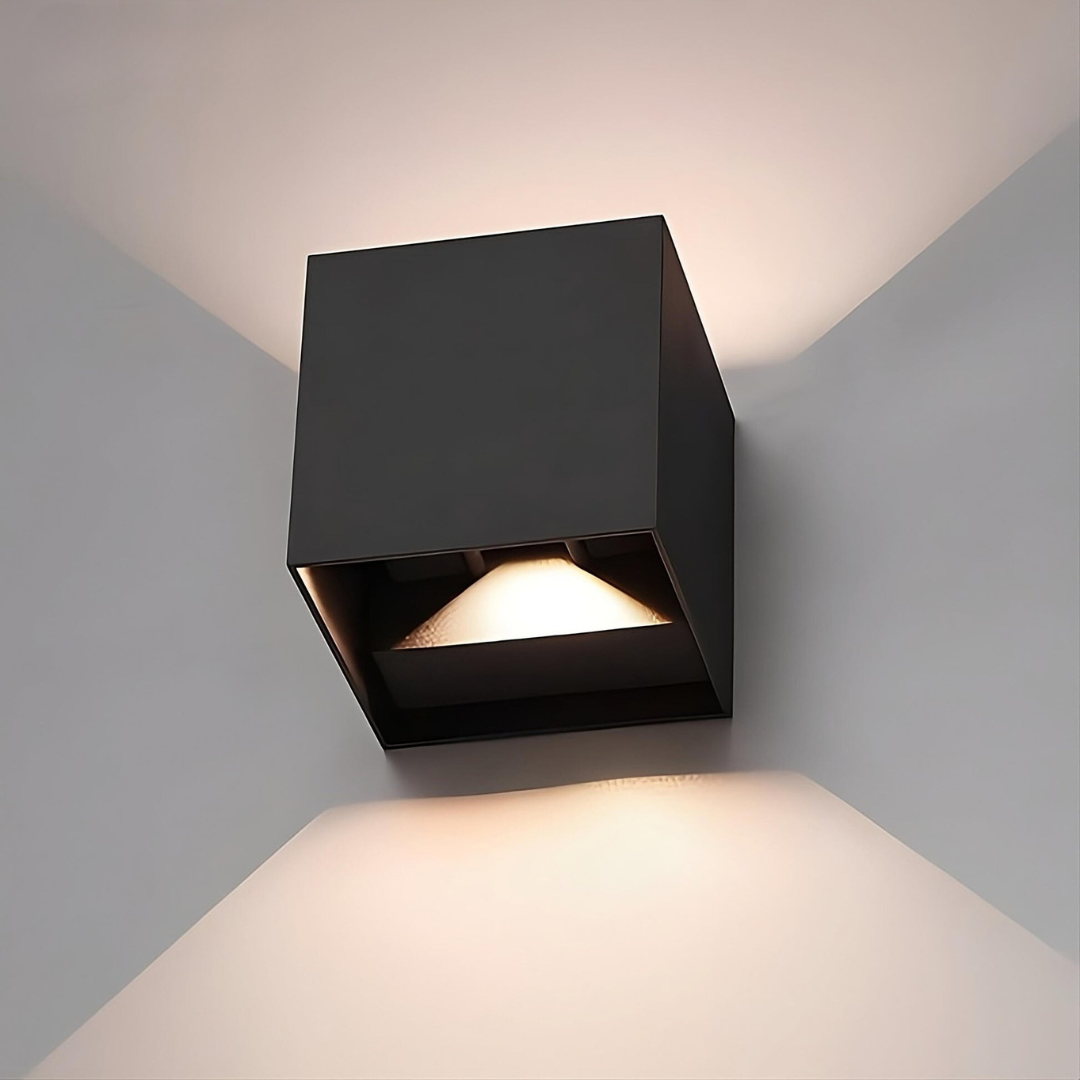 Close-up of a black cube wall lamp showing the open lower edge with its angled light diffuser, casting light up and down. 16