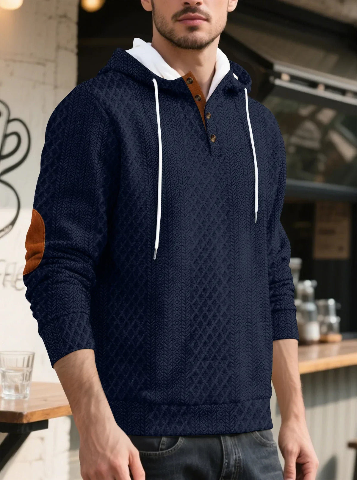 Model in a navy textured hoodie in a cafe setting, three-quarter view showing drawstrings, buttons and elbow patch 6