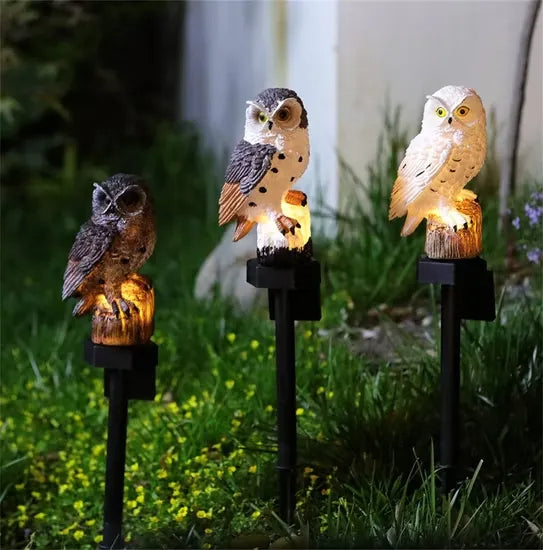 Three owl lights in brown, white-and-brown and white glowing on stakes in a garden bed with small yellow flowers. 8