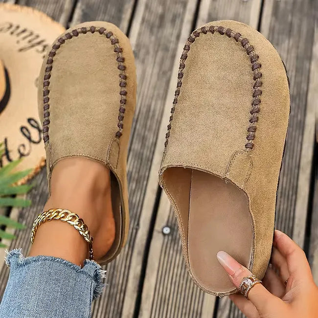 Light camel version of the stitched slip-on mules, one worn with a gold ankle chain, the other held up to show the footbed. 2
