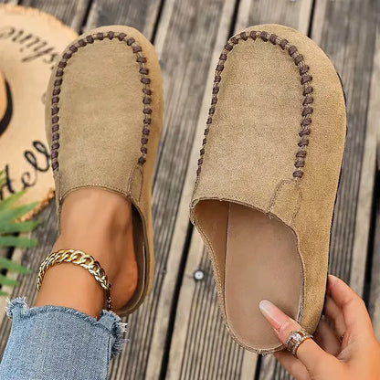 Light camel version of the stitched slip-on mules, one worn with a gold ankle chain, the other held up to show the footbed. 2