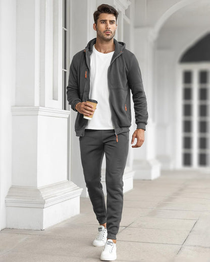 Model in dark grey tracksuit with jacket open over a white t-shirt, carrying a takeaway coffee outdoors 7