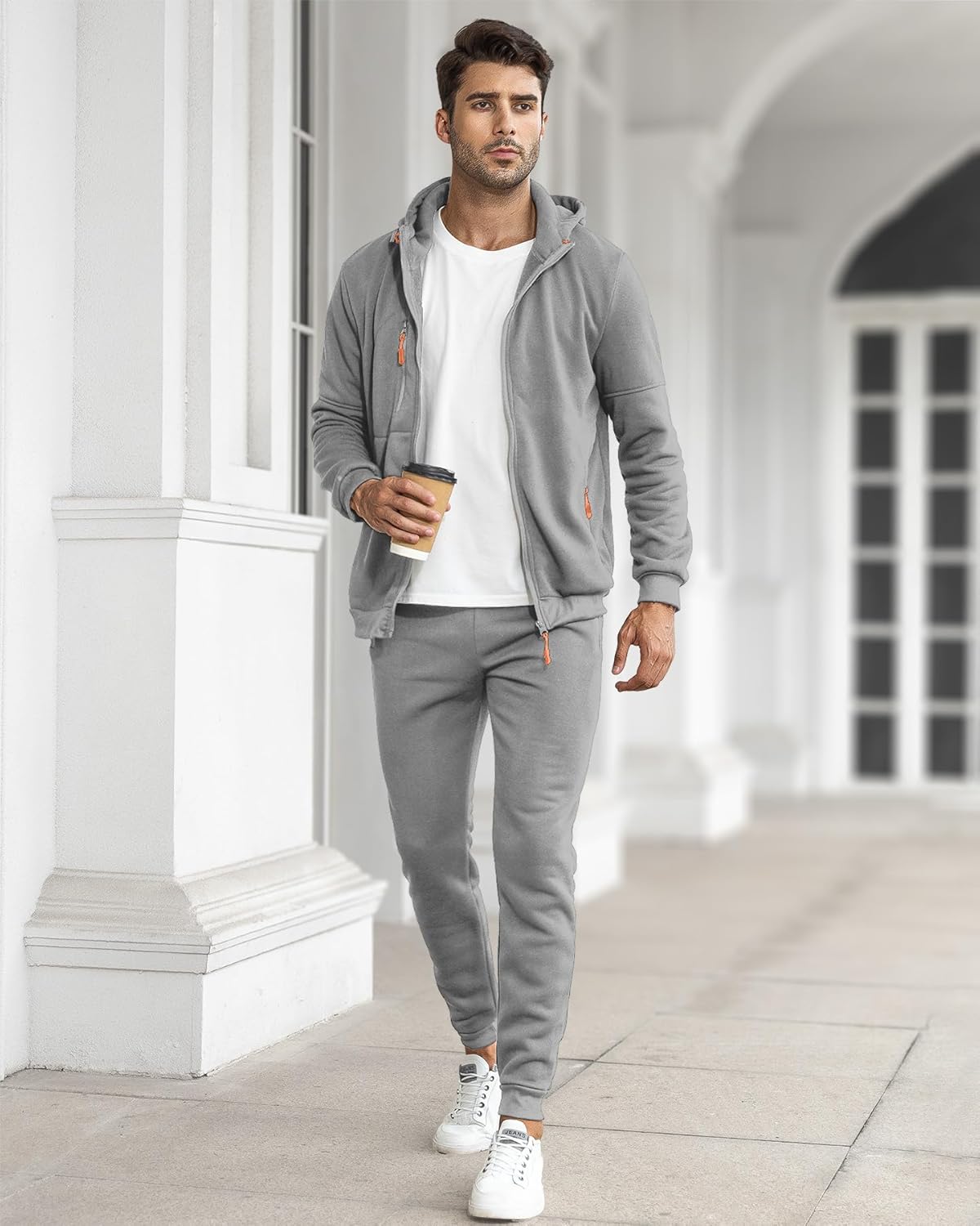 Man in the grey zip-up hooded tracksuit with orange zip pulls over a white T-shirt, walking along a pale colonnade 30