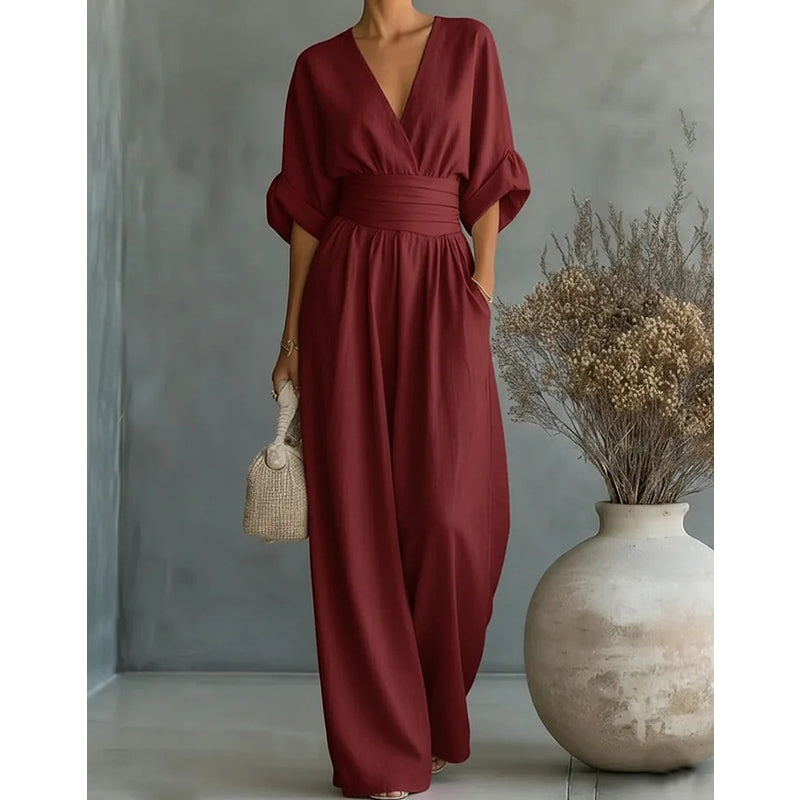 Burgundy jumpsuit worn by a model in a studio, showing the V-neck, ruched high waist and wide trousers beside a vase. 8