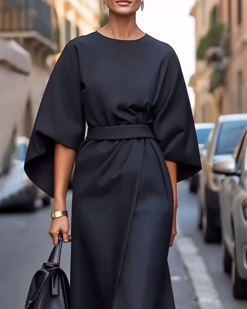 Close-up of the black dress's round neckline, wide sleeves and fabric belt, with the model wearing a gold cuff and holding a black bag. 5