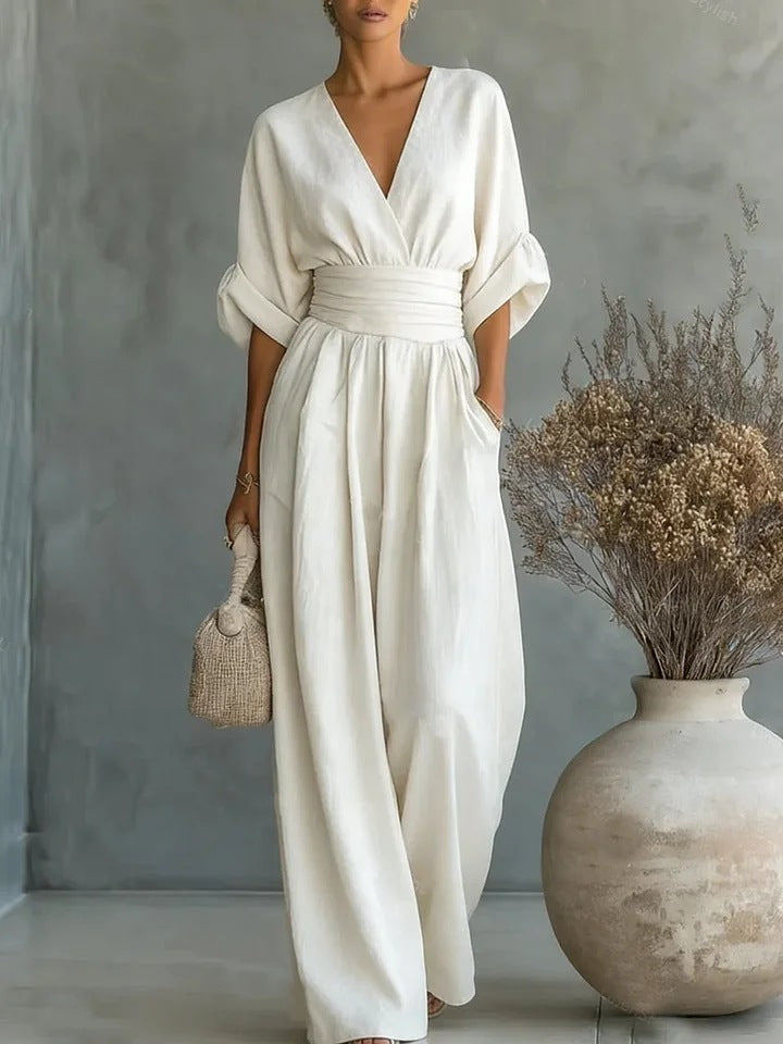 Model in the cream-white jumpsuit with wrapped V-neck, gathered waistband and wide legs, standing beside a large clay vase of dried flowers. 1