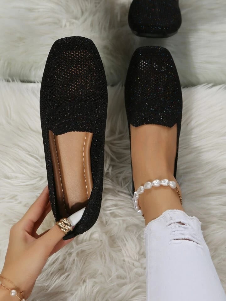 Black glittery mesh slip-on shoes on a fluffy white rug, one held up to show the brown footbed. 1