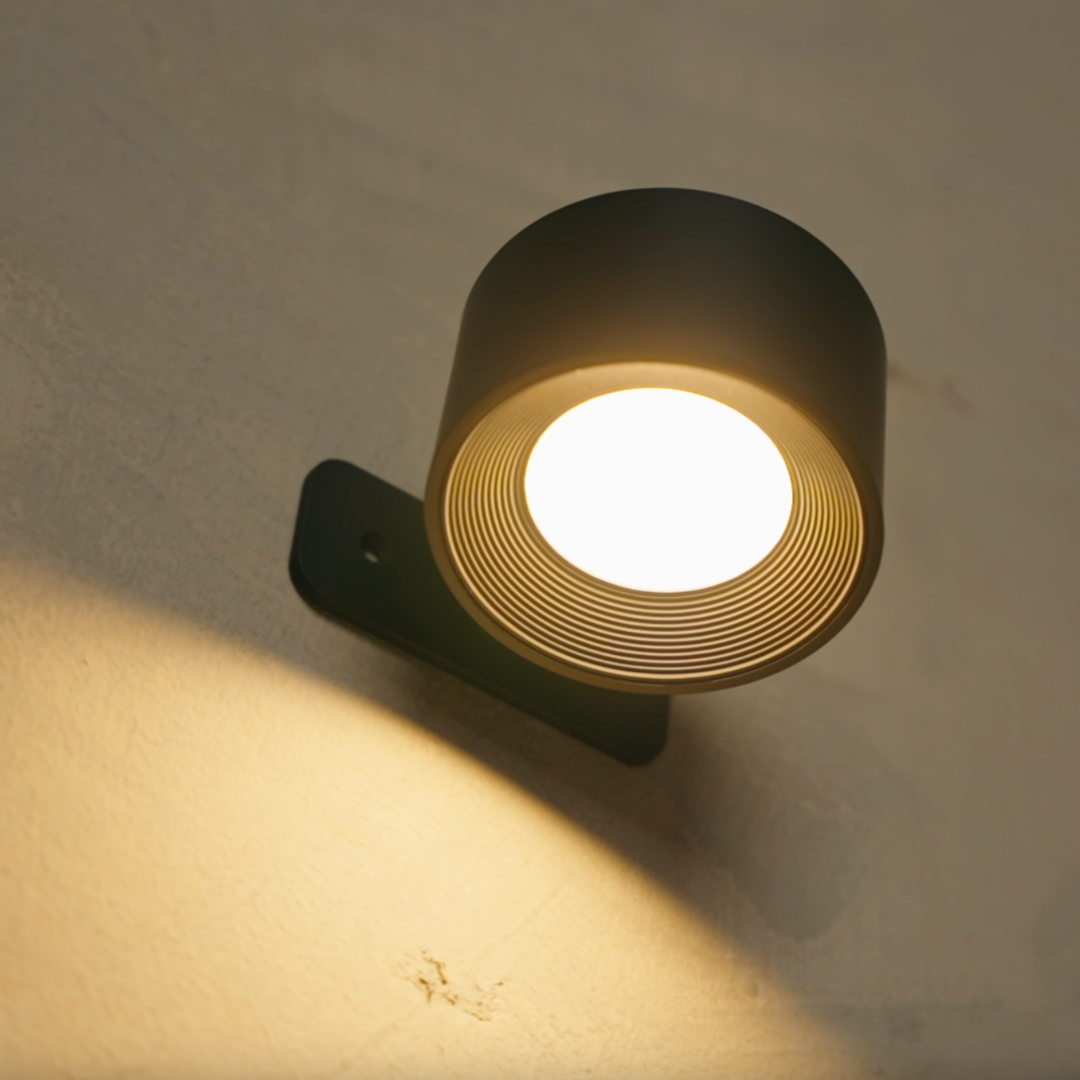 Close-up from below of the black wall light switched on, showing the ribbed inner ring, bright diffuser and mounting plate. 16