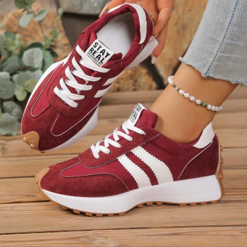 Burgundy suede trainers with white stripes, worn with rolled jeans and a pearl anklet on wooden decking 4