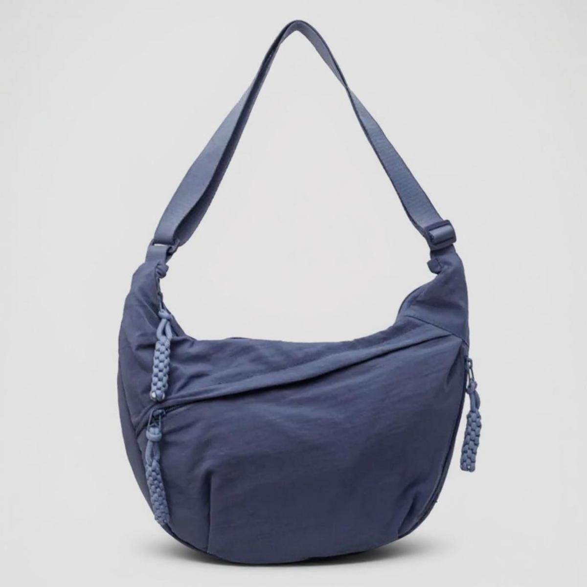 Front view of a crescent-shaped blue nylon bag with a single adjustable strap and braided zip pulls, on a plain grey background. 1