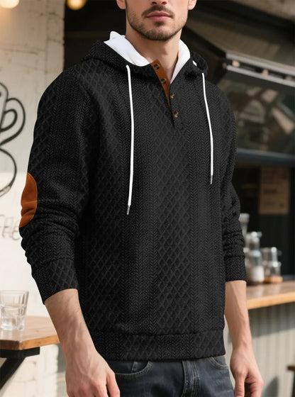 Model in a black textured hoodie in a cafe setting, three-quarter view showing buttons, drawstrings and tan elbow patch 7