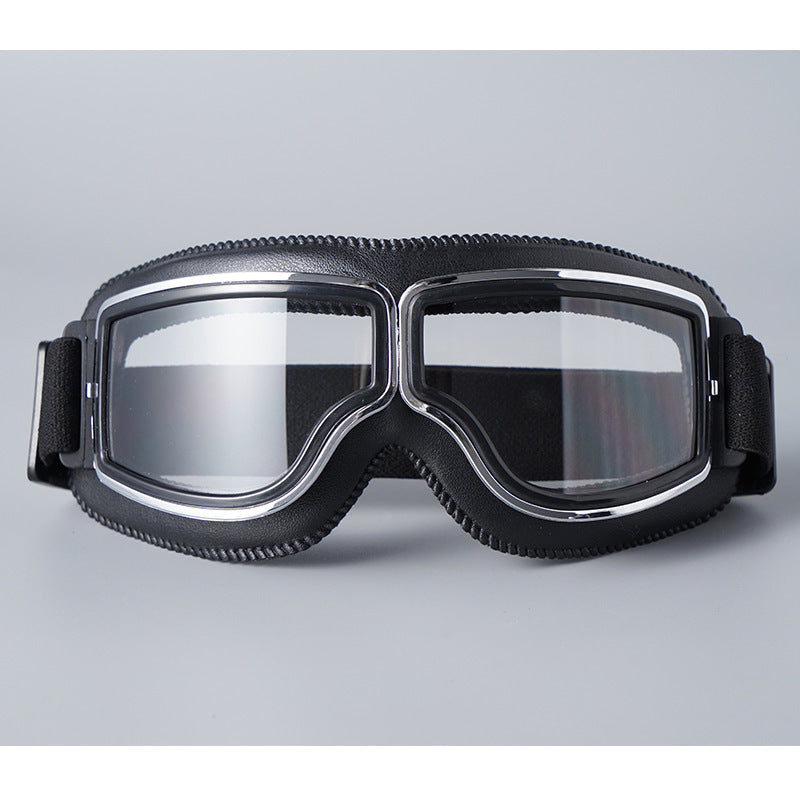 Front view of the black-padded goggles with clear lenses, chrome trim and a black elastic strap on a grey background. 16